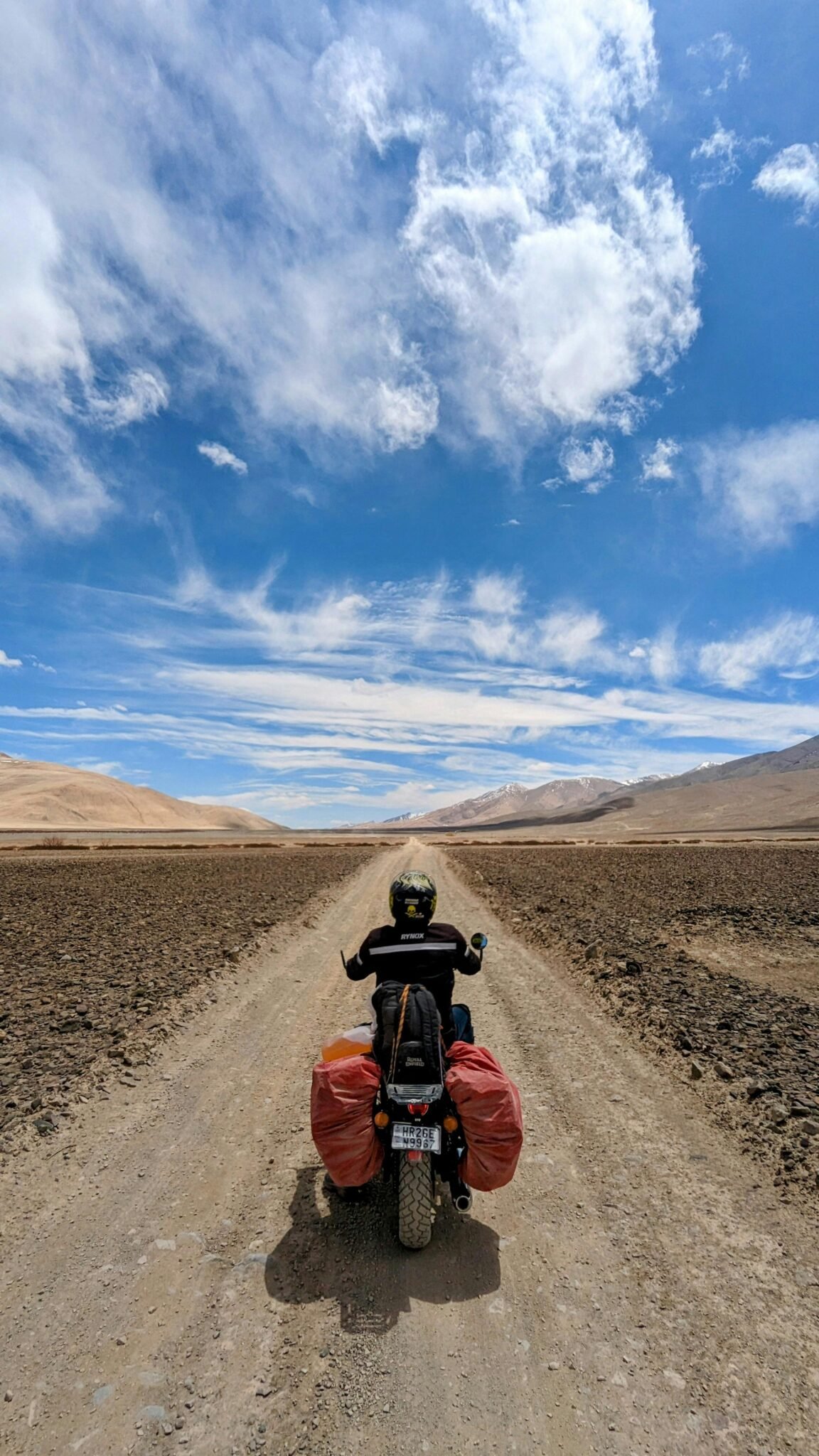 Motorcyclist rides down a dirt road in a vast desert landscape, under a bright blue sky.