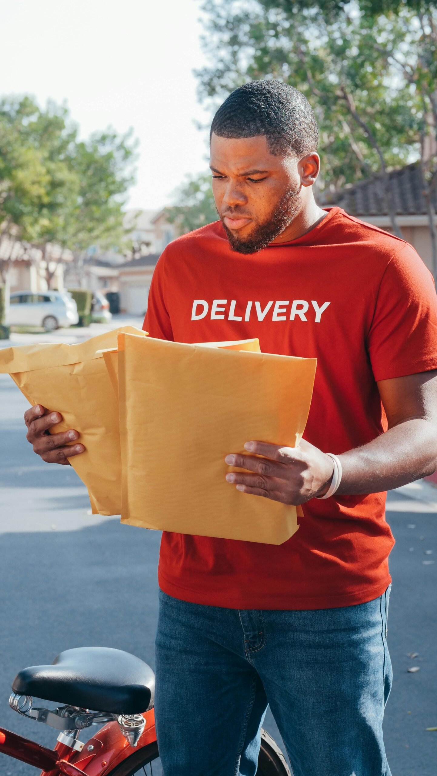 A delivery man in a red uniform holds envelopes while standing outdoors.